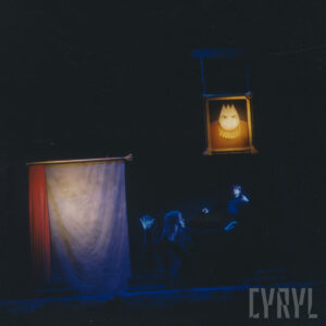 z lewej oświetlony płat materiału, w głębi dwoje aktorów, nad nimi wisi oświetlony portret lalki