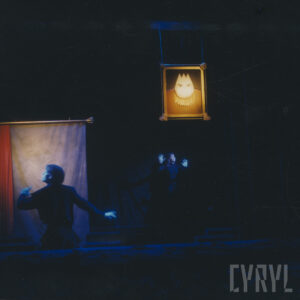 na pierwszym planie, z lewej mężczyzna odwrócony tyłem, za nim płat materiału, w głębi kobieta z uniesionymi dłońmi, nad nią zawieszony na linach i oświetlony portret lalki