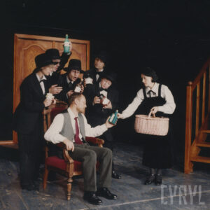 pośrodku na fotelu siedzi mężczyzna w ręce trzyma butelkę, z prawej strony stoi kobieta z koszykiem w ręku, za fotelem grupa kobiet i mężczyzn w czarnych garniturach i w kapeluszach