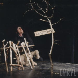 lalka osiołka siedzi w prowizorycznym szałasie z gałązek, obok małe, suche drzewko z przymocowanym do niego napisem Kłapouchy napisanym z błędami, przy lalce siedzi aktor ją animujący