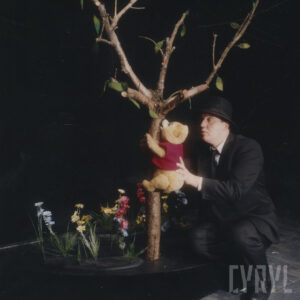 scenografia z małym drzewkiem i kwiatami koło niego, po drzewie wspina się lalka Kubusia Puchatka, podtrzymuje ją aktor w czarnym garniturze i kapeluszu na głowie