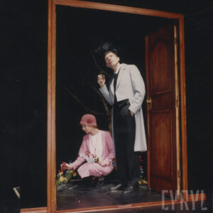 scenografia teatralna stworzona z otwartych drzwi, za nimi stoi mężczyzna, obok niego w przyklęku kobieta dotyka ręką kwiatów