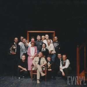 zdjęcie grupowe, pośrodku siedzi mężczyzna w fotelu, wokół niego kilkanaście osób, część z nich trzyma w rękach lalki