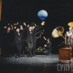 w głębi grupa aktorów w czarnych garniturach trzyma w rękach uniesionych nad głowami czarne kapelusze, po prawej stronie przykucnął jeden z aktorów, w rękach trzyma sznurek na końcu którego przywiązano niebieski balonik, do którego przymocowano lalkę Kubusia Puchatka, z prawej stoi mężczyzna z rozłożonym żółtym parasolem, na pierwszym planie gramofon z tubą