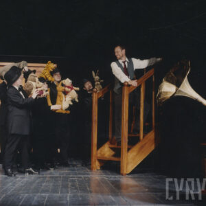 pośrodku sceny fragment schodów z balustradami po obu stronach, na nich stoi mężczyzna, z lewej grupa aktorów w czarnych garniturach i kapeluszach trzyma w rękach lalki, bohaterów spektaklu, z prawej tuba gramofonu