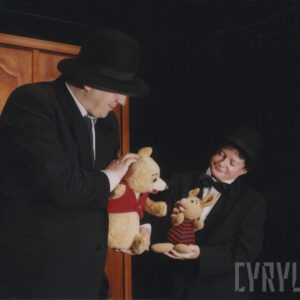 dwoje aktorów w czarnych garniturach i kapeluszach animuje lalki, z lewej mężczyzna trzyma lalkę Kubusia Puchatka, z prawej kobieta z Prosiaczkiem