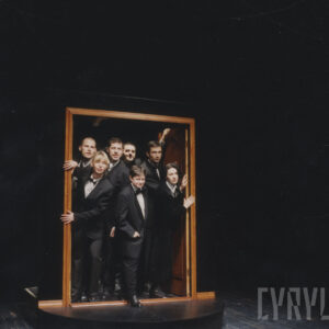 element scenografii w formie ościeżnica z otwartymi drzwiami, w niej grupa kobiet i mężczyzn w czarnych garniturach