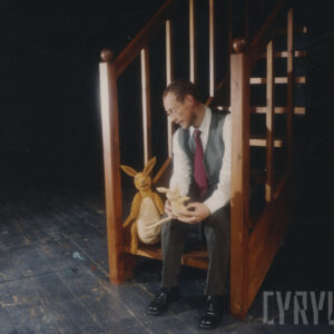 na drewnianych schodach z balustradą po obu stronach siedzi mężczyzna, obok niego lalka Królika