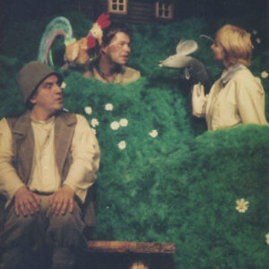 scenografia spektaklu z zieloną trawą i drewnianym domkiem na szczycie, między scenografią dwójka aktorów z lalkami kogucika i myszki, poniżej mężczyzna w kostiumie siedzi na niskiej drewnianej ławce