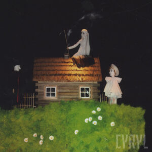 scenografia spektaklu z zieloną trawą i drewnianym domkiem, po lewej i prawej stronie dwie lalki, na dachu domku lalka symbolizująca śmierć