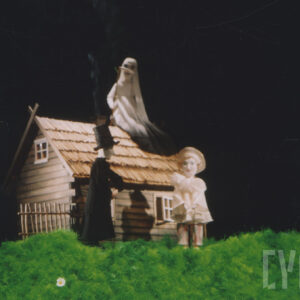 scenografia spektaklu z zieloną trawą i drewnianym domkiem, przed nim dwie lalki, na dachu domku lalka symbolizująca śmierć