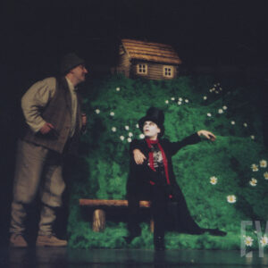 scenografia spektaklu z zieloną trawą i drewnianym domkiem na szczycie, poniżej dwóch aktorów w kostiumach, jeden z nich siedzi na drewnianej ławce