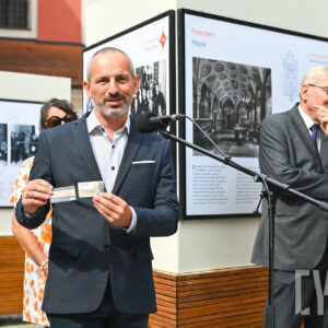 mężczyzna w garniturze przemawia do mikrofonu i prezentuje kartę tramwajową, obok niego mężczyzna i kobieta, w tle fragment wystawy wydruków wielkoformatowych na stelażach