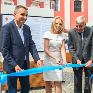 dwaj mężczyźni i kobieta przecinają wstęgę na tle wielkoformatowego wydruku umieszczonego na stelażu, w głębi fragment budynku