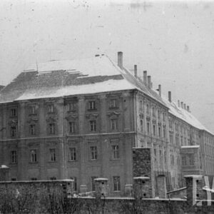 nieotynkowany, kilkupiętrowy budynek z rzędami okien i spadzistym dachem, w głębi fragment dzwonnicy