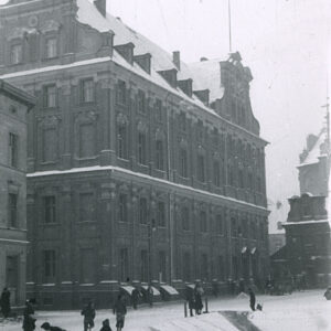 duży, kilkupiętrowy budynek z rzędami okien i ozdobnymi szczytami w zimowej scenerii, przed budynkiem plac ze ślizgawką dla dzieci we wgłębieniu terenu