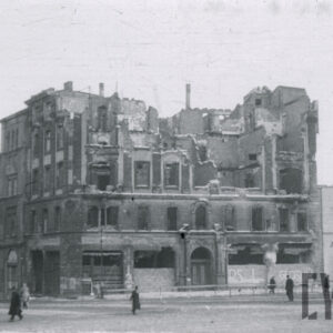 zrujnowany, pozbawiony górnych kondygnacji narożnikowy budynek hotelu, na pierwszym planie fragment ulicy z przechodzącymi ludźmi