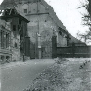 zniszczona drewniana dzwonnica i fasada kościoła z blendami i ozdobnym szczytem, obok dzwonnicy fragment murowanego ogrodzenia z kutą bramą