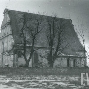 budynek kościoła z wysokim, dwuspadowym dachem krytym dachówką i ozdobnym szczytem, budynek przysłaniają nagie drzewa, na pierwszym planie otaczający teren kościelny ceglany mur