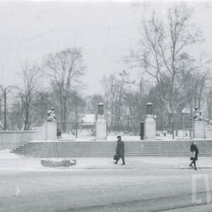 murowane ogrodzenie z kutą bramą poprzedzone szerokimi schodami w zimowej scenerii, na pierwszym planie ulica i nieliczni przechodnie