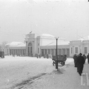 plac przed piętrowym budynkiem dworcowym z centralnie umieszczonym wykuszem, na placu wozy konne, na placu i dachu budynku leży śnieg