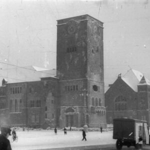 potężna, nieregularna bryła zamku z ośnieżonymi dachami i płasko zakończoną wieżą, na pierwszym planie przechodnie i ciężarówka