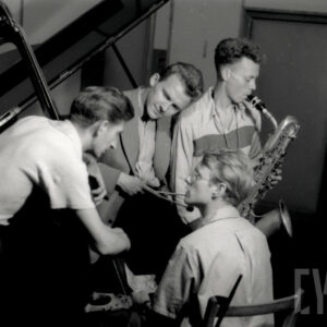 kilku młodych mężczyzn z instrumentami, jeden z nich siedzi przy fortepianie, w studio nagraniowym