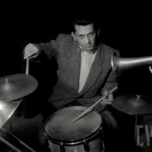 młody mężczyzna w marynarce w kratę siedzi i gra na perkusji, po jego obu stronach widać fragmenty trąbki i puzonu