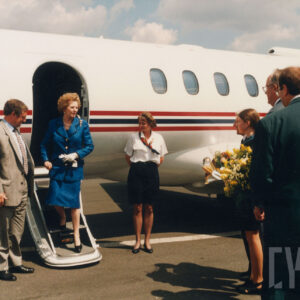 grupa osób stojąca stojąca przed samolotem, czekająca na wysiadającą z niego starszą, elegancko ubraną kobietę