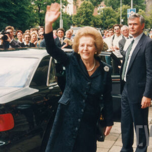 starsza elegancko ubrana kobieta pozdrawiająca ludzi zgromadzonych wokół samochodu, którym przyjechała