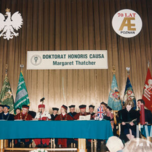 grupa osób w togach rektorskich zasiadających przy stole prezydialnym, za nimi sztandary