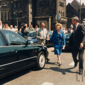 elegancka, starsza kobieta zmierzająca do samochodu, w tle widoczny duży budynek z piaskowca i grupa gapiów