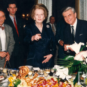 grupa elegancko ubranych, starszych osób, jedna kobieta i dwóch mężczyzn, pozujących do fotografii przy zastawionym stole