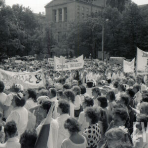 tłum ludzi na placu, większość w białych kitlach, część trzyma transparenty z hasłami, w głębi duży budynek 