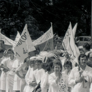 duża grupa kobiet, między nimi kilku mężczyzn, większość w białych kitlach, część kobiet ma na głowach pielęgniarskie czepki, część trzyma nad głowami transparenty z hasłami