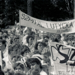 duża grupa kobiet, między nimi kilku mężczyzn, większość w białych kitlach, część kobiet ma na głowach pielęgniarskie czepki, część trzyma nad głowami transparenty z hasłami