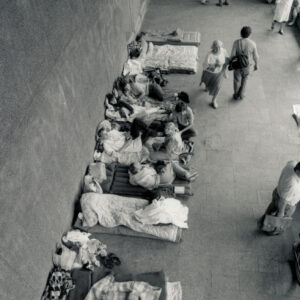 widok z góry na ludzi leżących lub siedzących na materacach pod ścianą budynku, u góry stoi grupa ludzi