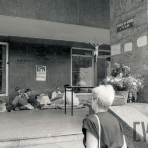 kilka osób siedzi lub leży na materacach pod ścianą budynku, z prawej na kolumnie i na szybie naklejone transparenty z hasłami, z prawej krzyż z figurą Chrystusa