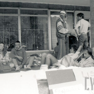 kilka osób siedzi na materacach pod ścianą budynku i przy kamiennym słupie budynku, z prawej stoi trzech mężczyzn