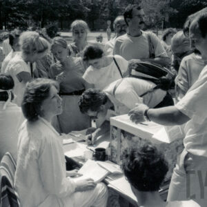 kobieta i mężczyzna siedzą przy stoliku, na nim stoi przeźroczysta urna z pieniędzmi, wokół stolika ludzie, jeden mężczyzna pochylony nad stolikiem coś pisze lub podpisuje