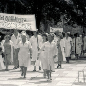 grupa kobiet i kilku mężczyzn w białych kitlach idzie chodnikiem, część z kobiet w czepkach pielęgniarskich, z przodu dwie kobiety niosą duży transparent