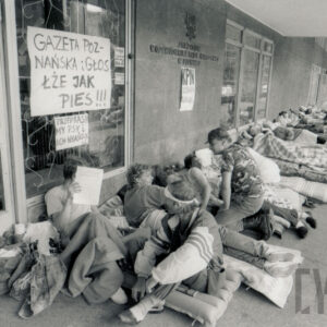 grupa ludzi leżąca lub siedząca na materacach przy ścianie budynku, nad szybie transparenty z hasłami