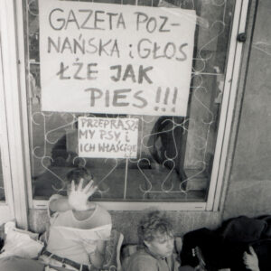 dwóch młodych mężczyzn leży na materacach pod ścianą budynku, jeden z nich, z lewej zasłania swoją twarz wyciągniętą przed siebie dłonią, za nimi okno, na nim naklejone transparenty z hasłami krytykującymi prasę