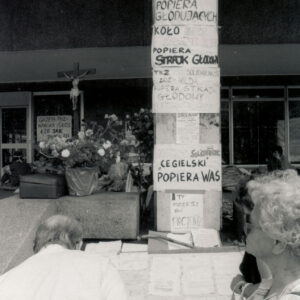 transparenty z hasłami zawieszone na kamiennym słupie budynku, z lewej kwiaty w pojemniku, za nimi krzyż z figurą Chrystusa