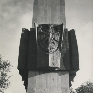 prosta, pionowa, gładka, wysoka, prostokątna bryła, w połowie jej wysokości wiszą ciemne tablice z płaskorzeźbami przedstawiającymi sylwetki ludzi, przed bryłą stoi brązowa rzeźba dwóch żołnierzy w wojskowych płaszczach i rogatywkach na głowach, w dłoniach trzymają broń, jeden karabin, drugi opuszczoną szablę