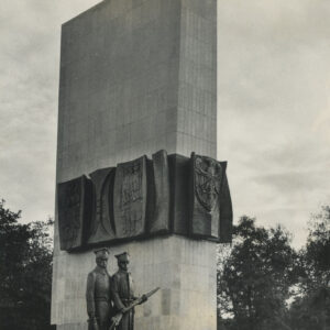 prosta, pionowa, gładka, wysoka, prostokątna bryła, w połowie jej wysokości wiszą ciemne tablice z płaskorzeźbami przedstawiającymi sylwetki ludzi, przed bryłą stoi brązowa rzeźba dwóch żołnierzy w wojskowych płaszczach i rogatywkach na głowach, w dłoniach trzymają broń, jeden karabin, drugi opuszczoną szablę
