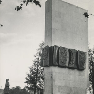 prosta, pionowa, gładka, wysoka, prostokątna bryła, w połowie jej wysokości wiszą ciemne tablice z płaskorzeźbami przedstawiającymi sylwetki ludzi, przed bryłą stoi brązowa rzeźba dwóch żołnierzy w wojskowych płaszczach i rogatywkach na głowach, w dłoniach trzymają broń, jeden karabin, drugi opuszczoną szablę