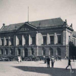 monumentalny, klasycystyczny budynek z kolumnowym wejściem i dekoracyjnym frontonem; przed budynkiem stoją stare samochody, a na chodniku przechodzą ludzie