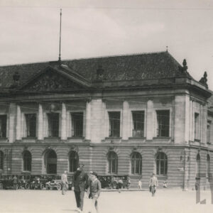 monumentalny, klasycystyczny budynek z kolumnowym wejściem i dekoracyjnym frontonem; przed budynkiem stoją stare samochody, a na chodniku przechodzą ludzie