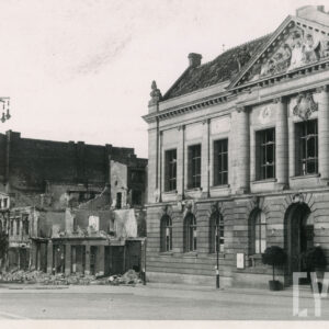 monumentalny, neorenesansowy budynek z niedużymi uszkodzeniami i wybitymi oknami, sąsiadujący ze zrujnowaną kamienicą, na chodniku widoczny gruz, obok wysoka, ozdobna latarnia